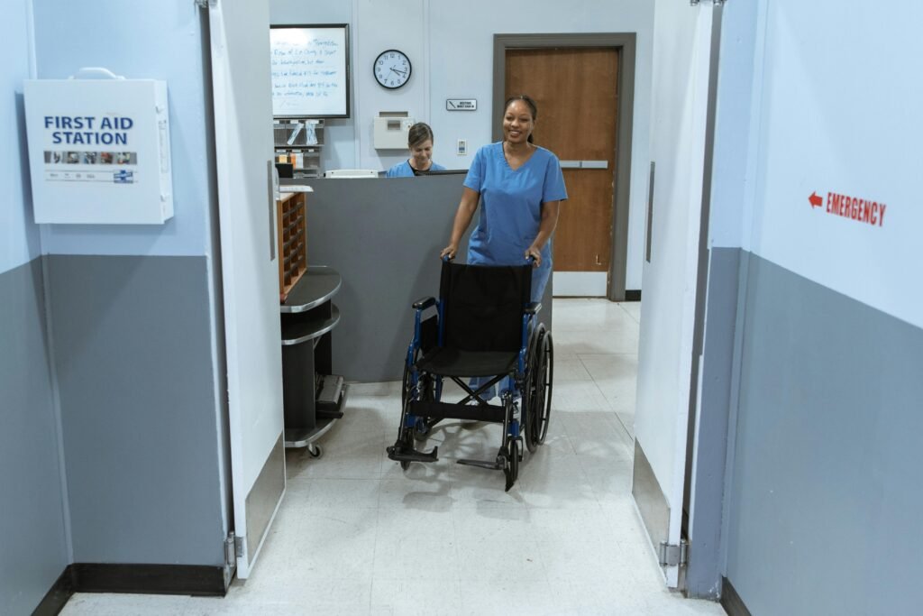 A nurse in scrubs pushes an empty wheelchair through a hospital hallway.
